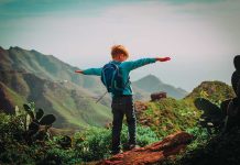 Child with backpack stands on mountain arms outstretched