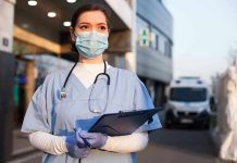 Nurse in scrubs and mask outside hospital holding clipboard