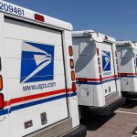 Row of USPS mail trucks parked in lot.