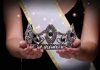 Sash-wearing person holding an ornate jeweled crown.