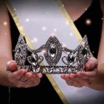 Sash-wearing person holding an ornate jeweled crown.