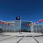 Flags outside NATO headquarters building under clear blue sky.