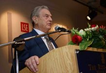 A man in a suit speaking at a podium with flowers and a microphone