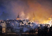 A large crowd of police officers in helmets facing a smoky protest scene at night