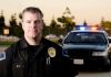 A police officer in uniform stands in front of a police car with lights on