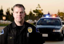 A police officer in uniform stands in front of a police car with lights on