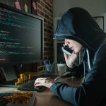 Massive Crypto Heist: What It Means for International Cybersecurity Efforts Hacker in hood using computer and phone analyzing code