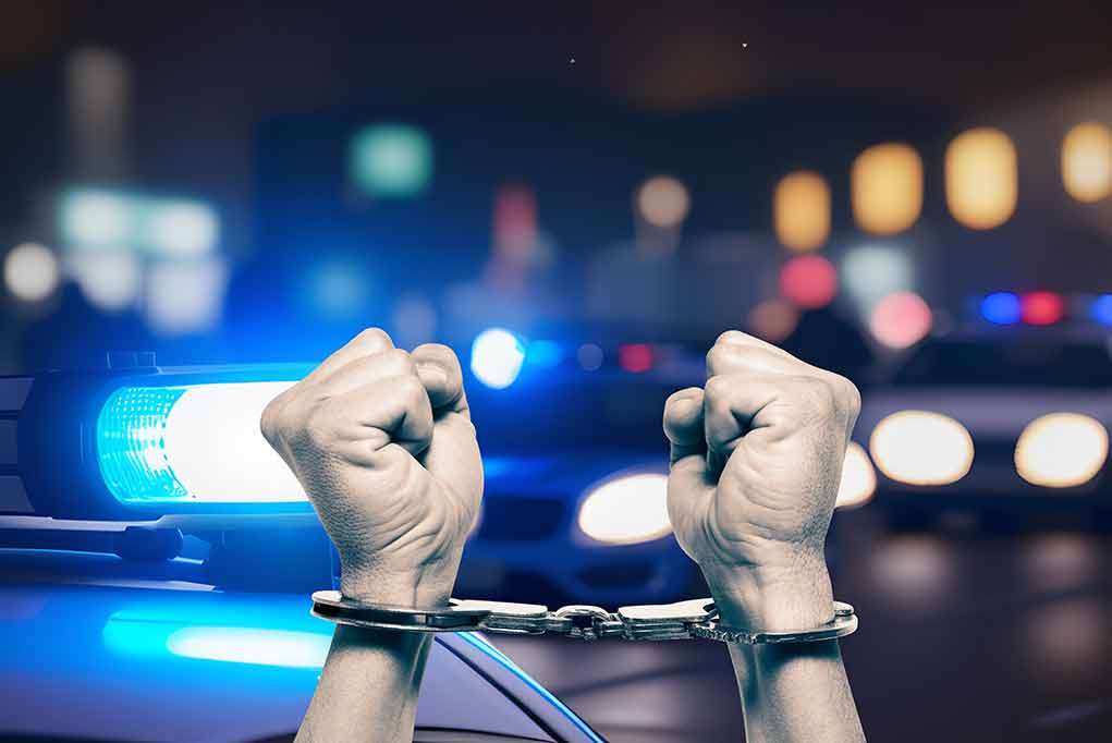 2348949967 featured image Handcuffed hands raised, police cars with lights on.