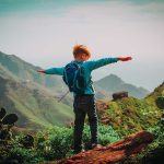 Child with backpack stands on mountain arms outstretched