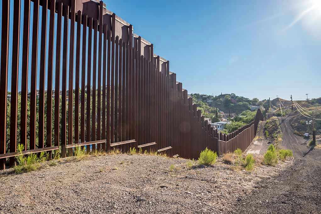 657897121 Tall metal border wall with rural landscape.