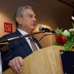 A man in a suit speaking at a podium with flowers and a microphone