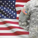 Hegseth Doubles Down — Combat Roles Under Fire! Soldier in camouflage uniform standing before American flag.