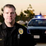 Off-Duty Cop Crashes Trump Event – Exploits Protocols! A police officer in uniform stands in front of a police car with lights on