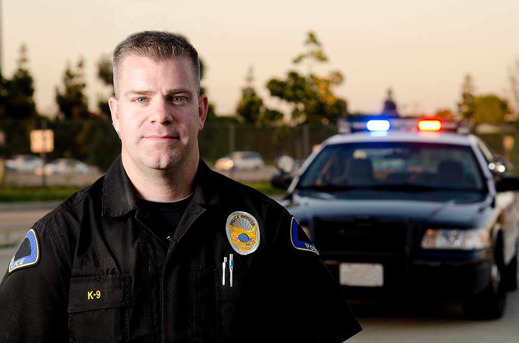 A police officer in uniform stands in front of a police car with lights on