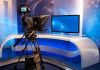 TOP News Anchor BUSTED – Massive Scam EXPOSED TV studio with camera and empty anchor desk.