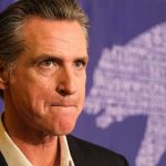 Newsom’s TOP Aide ARRESTED – Money Missing! Man in suit, solemn expression, purple background with bear drawing.
