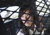 A woman sitting in the back of a police car, looking through a protective mesh barrier