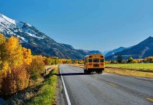A yellow school bus driving on a rural road surrounded by autumn trees and mountains