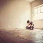 A child sitting alone in an empty room, covering their face with their hands