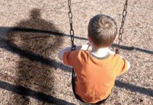 A young boy sitting on a swing at a playground, with his shadow cast on the ground