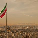 Trump’s Iran Blitz—Military Decimated in Weeks Iranian flag waving over a city skyline with mountains in the background