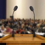 100-Year-Old Warning Haunts Modern Economics A wooden podium with two microphones in front of a blurred audience