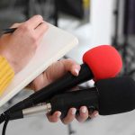 America’s Visa Crackdown Leaves Canada Facing 600-Day Waits Journalist holding two microphones while writing in a notebook