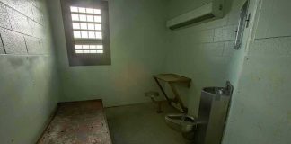 Empty jail cell with bed, barred window, and stainless steel toilet