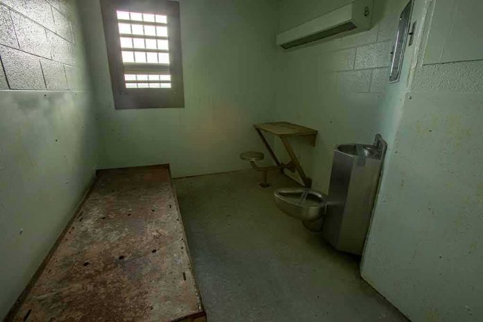 Empty jail cell with bed, barred window, and stainless steel toilet