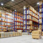 Retail Warehouses Become War Targets Warehouse aisles with tall blue and orange pallet racks filled with boxes