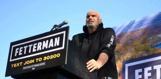 Dem Base Revolts: Fetterman Slide Speaker at outdoor rally standing at podium with microphone