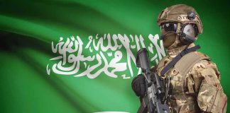 Soldier in tactical gear stands before a green flag