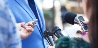 Reporters hold microphones toward a person in a blue suit