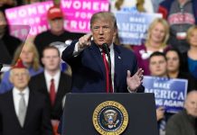 Donald Trump speaking at a rally podium, pointing toward the audience