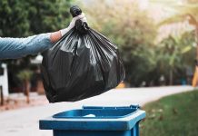 gloved hand holding a black trash bag over a blue garbage bin