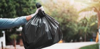 gloved hand holding a black trash bag over a blue garbage bin