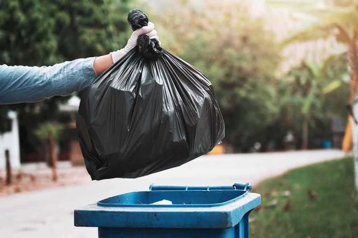 gloved hand holding a black trash bag over a blue garbage bin